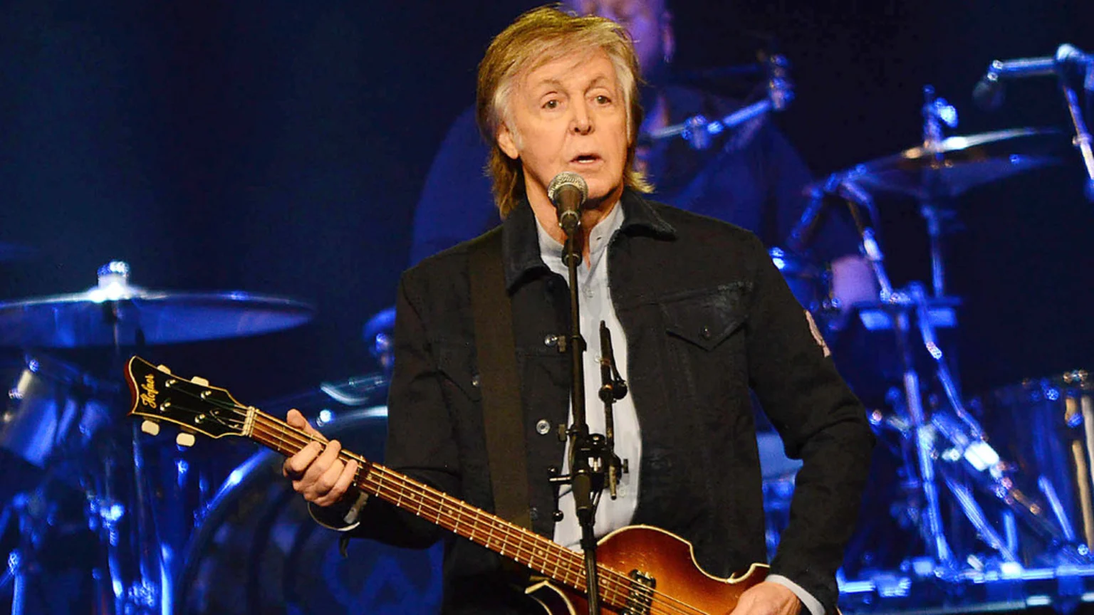 Paul McCartney performs live on stage, singing into a microphone while playing his iconic Hofner violin bass guitar, against a background of blue stage lights and blurred drum kit.