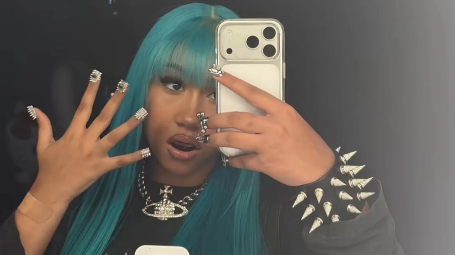 A person with teal hair and silver spiky nails takes a selfie, showing off a spiked bracelet, an orb necklace, and a bandage on their wrist.