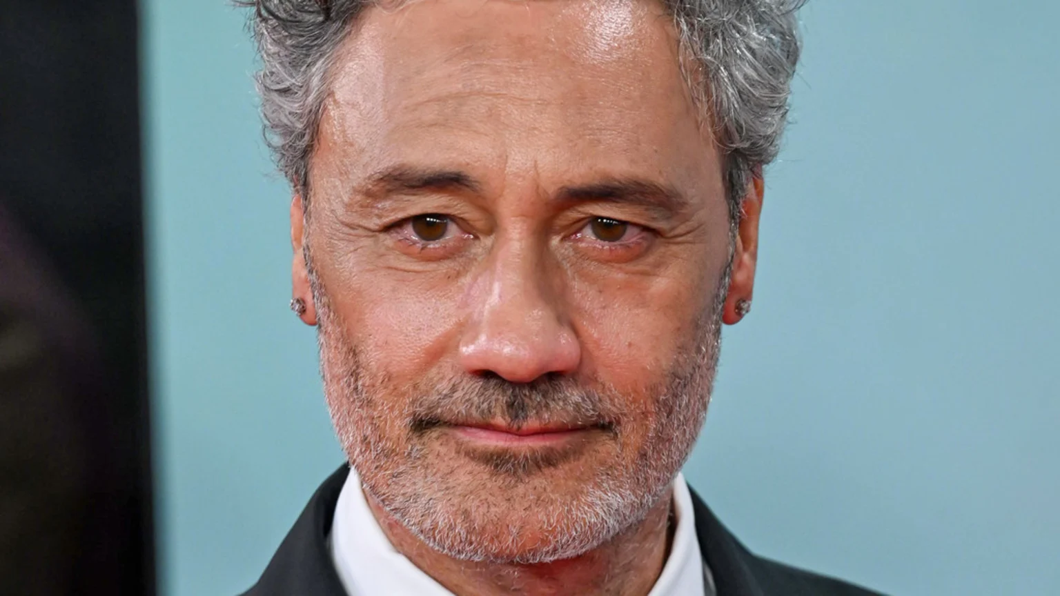 Close-up of Taika Waititi, featuring his grey, wavy hair, brown eyes, a greying beard, and stud earrings, wearing a dark suit, looking forward.