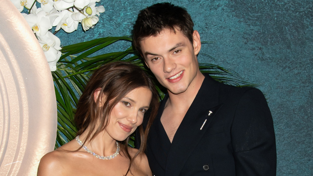 Millie Bobby Brown, in a diamond necklace, and Jake Bongiovi, in a dark jacket with a silver pin, smile at the camera against a floral and teal background.
