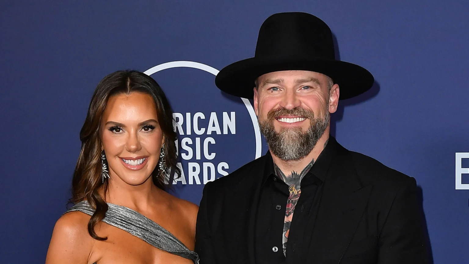 A smiling man with a beard, black hat, and tattoos on his neck stands next to a smiling woman with long brown hair, large earrings, and a metallic silver one-shoulder dress, posing in front of a "American Music Awards" backdrop.