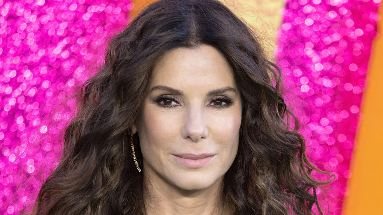 A close-up portrait of actress Sandra Bullock with dark wavy hair, wearing a long earring, looking directly at the camera with a slight smile against a blurred background of pink lights and orange.