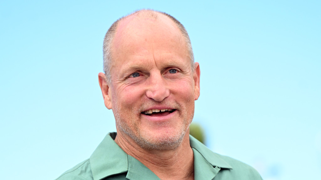 Woody Harrelson, with short hair and a light beard, smiles broadly with his mouth open, wearing a green shirt against a clear blue sky.