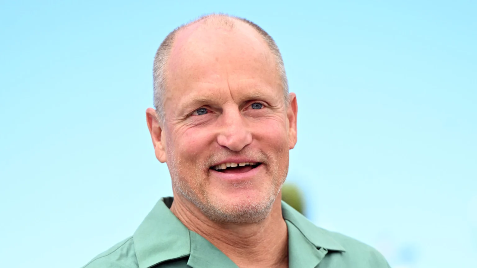 Woody Harrelson, with short hair and a light beard, smiles broadly with his mouth open, wearing a green shirt against a clear blue sky.