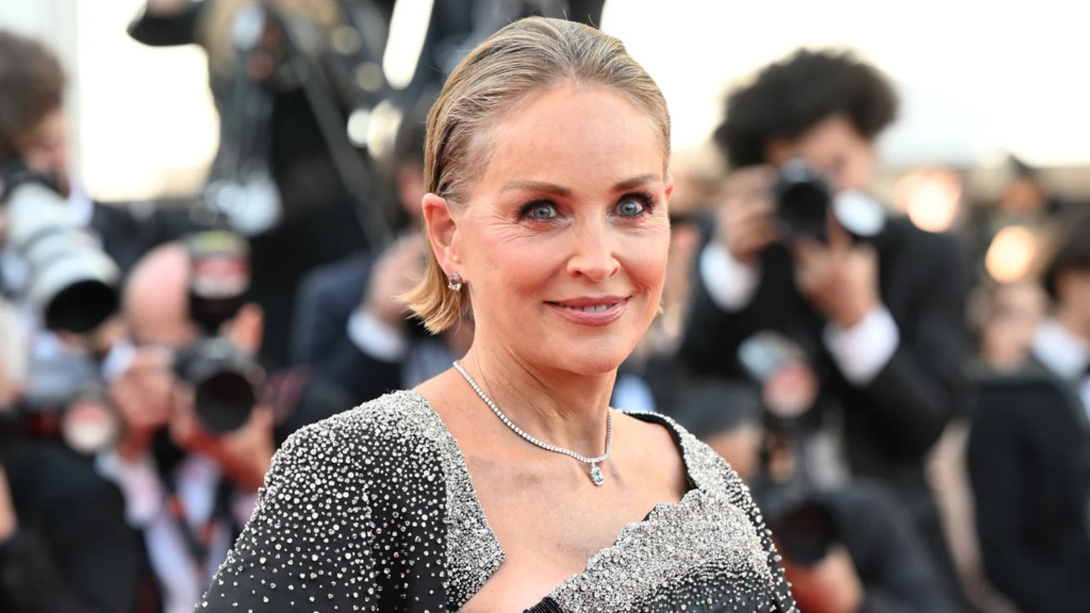 Sharon Stone smiles at the camera, wearing a black sparkling outfit and a diamond necklace, with a blurred crowd and photographers in the background.