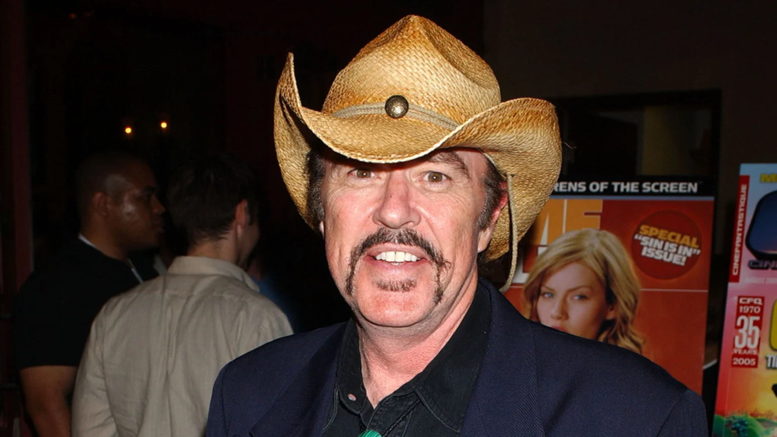 Close-up of a smiling man with a mustache and goatee, wearing a straw cowboy hat and a dark jacket, with blurred people and posters in the background.