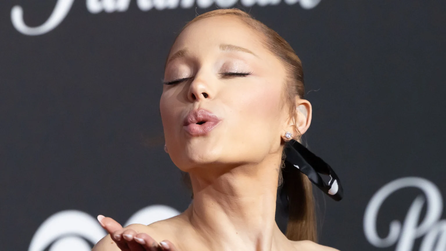 Ariana Grande, eyes closed, blowing a kiss, with a black ribbon in her hair, against a dark, blurred background.