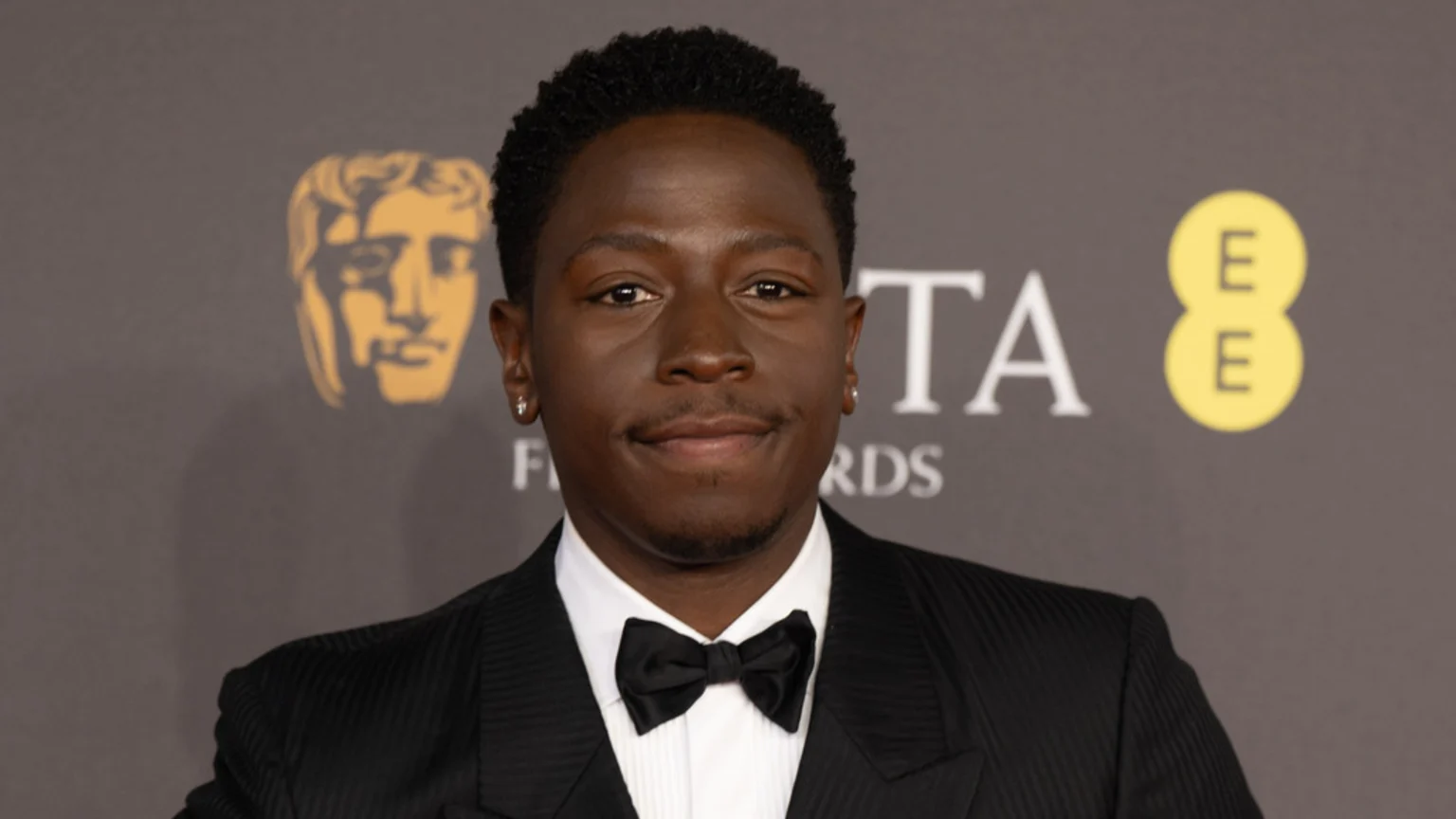 David Jonsson smiles at the camera, wearing a black tuxedo and bow tie, with the BAFTA mask and EE logos in the background.