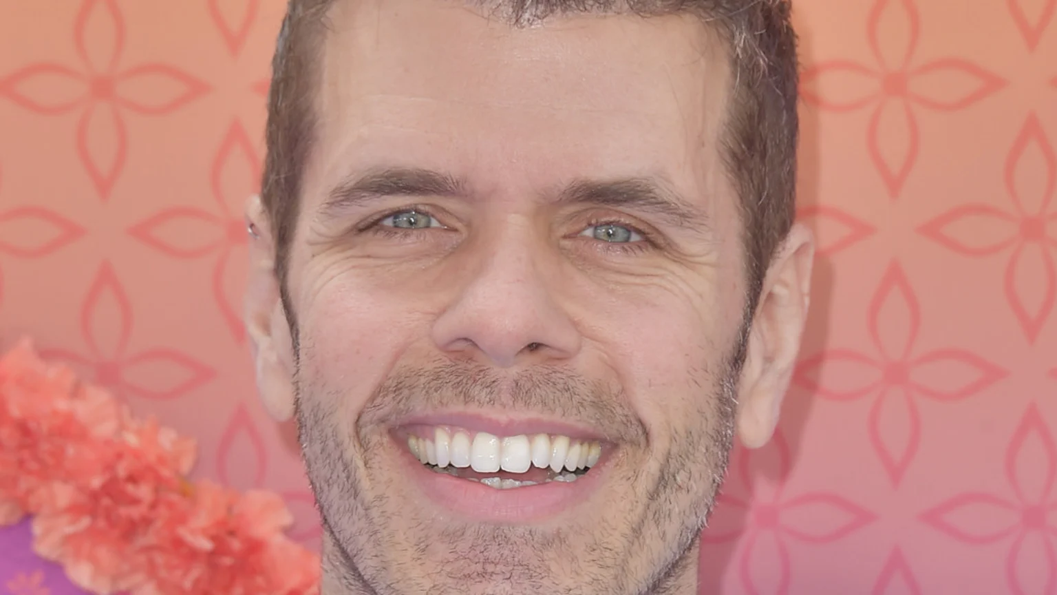 A smiling man with light blue eyes, light stubble, and an earring, shows his white teeth against a pink background featuring a repeating floral pattern and blurry orange flowers.