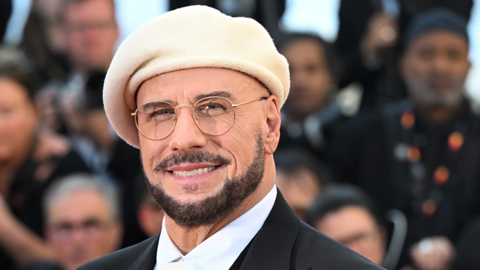 A smiling John Travolta wears a cream beret, round glasses, and a beard, with a blurred crowd in the background.
