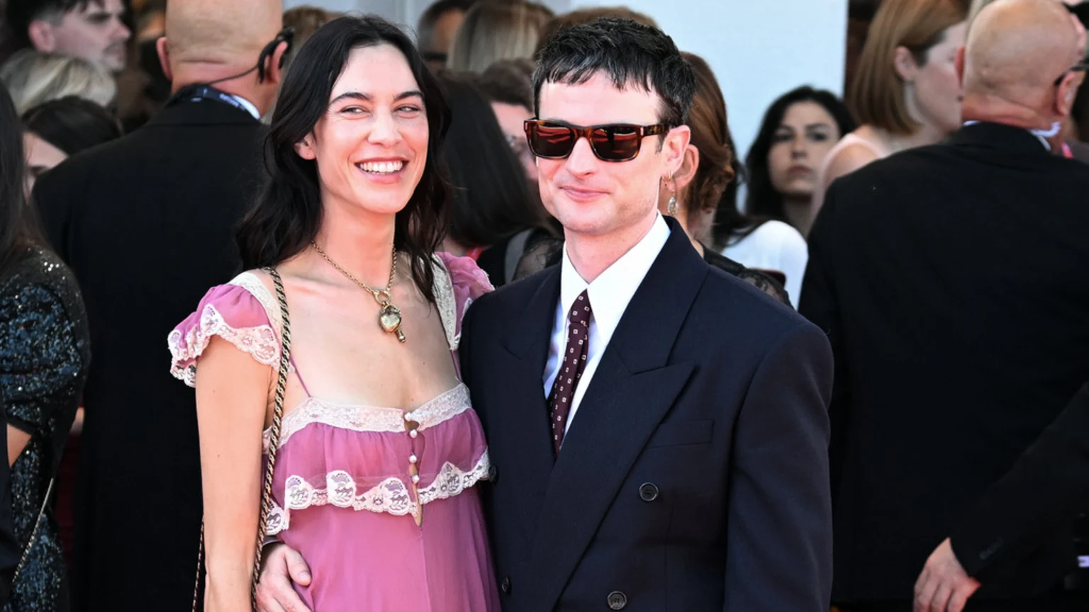 A broadly smiling woman in a pink dress with lace trim and a man in a dark suit, tie, and sunglasses with a subtle smile pose together, his hand on her waist, at an event.