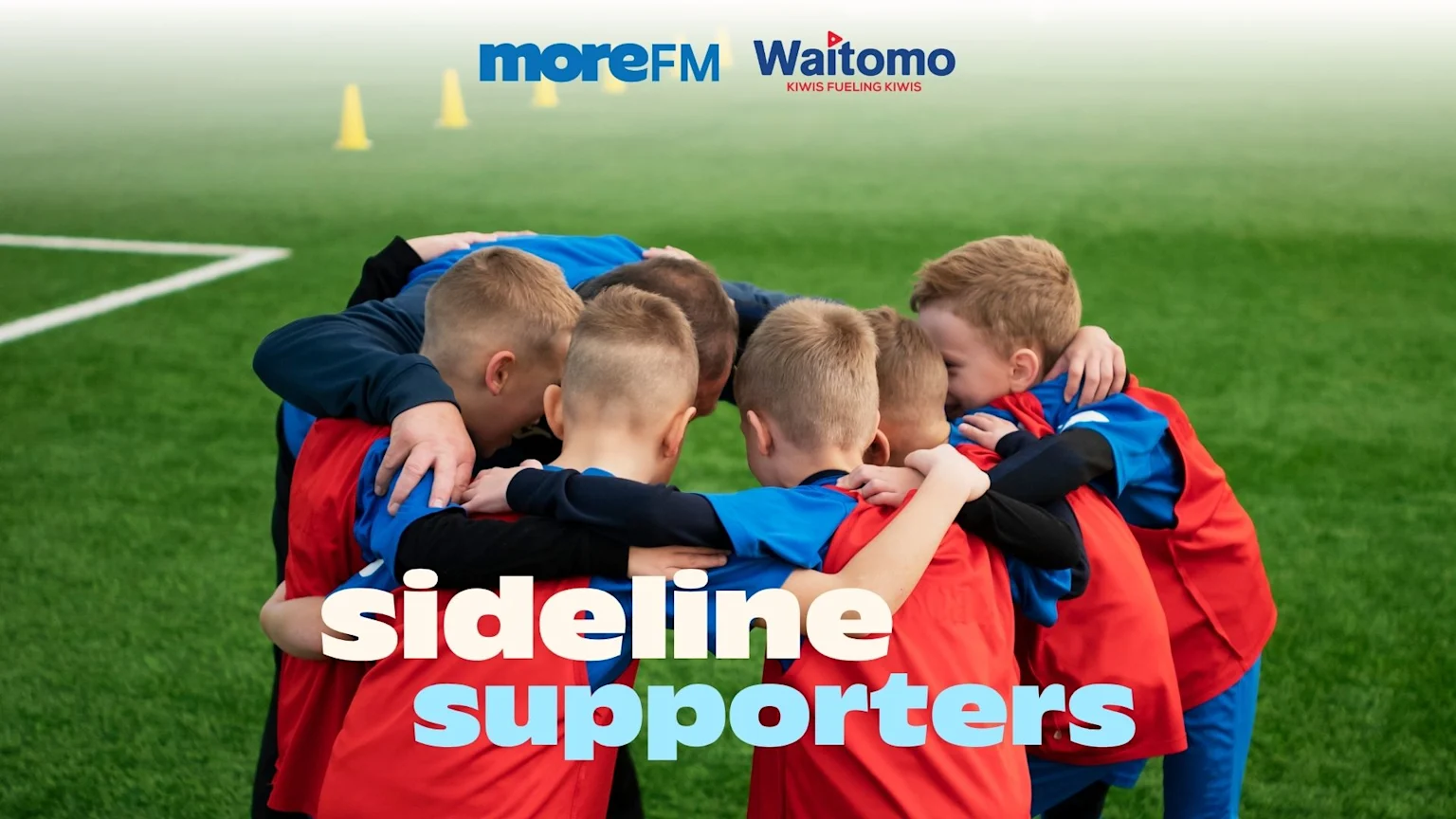 A group of young boys in red and blue jerseys huddle together on a green sports field, with "moreFM Waitomo" and "sideline supporters" text overlays.