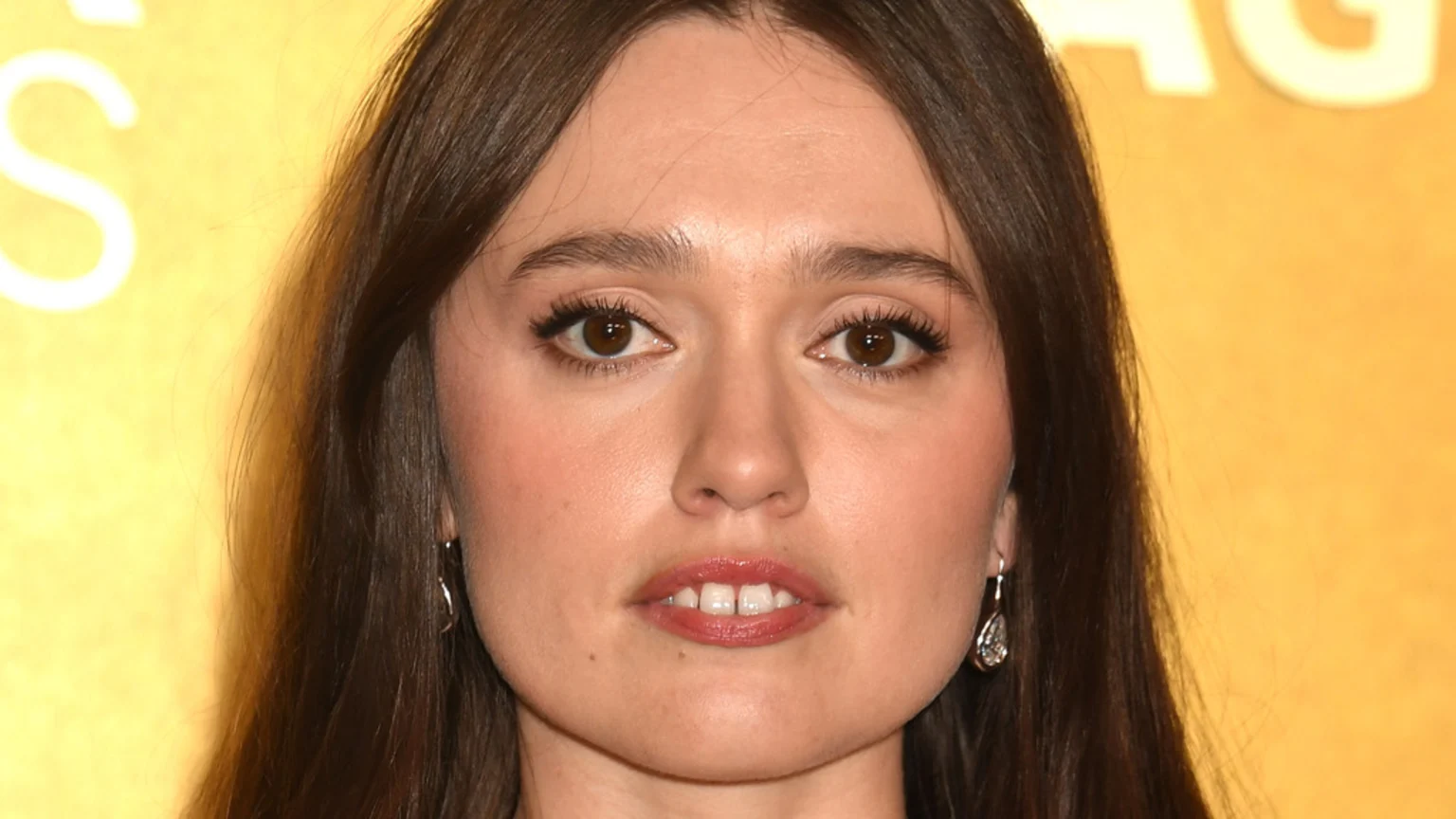 Close-up of a woman with long dark hair, brown eyes, and fair skin. Her lips are parted, revealing front teeth with a gap, and she wears silver drop earrings against a blurry yellow background.