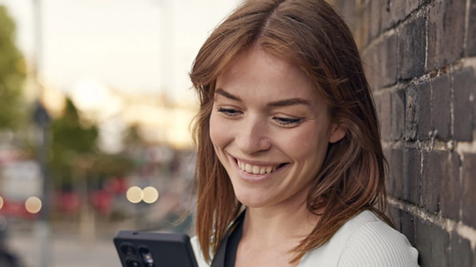 A smiling young woman with reddish-brown hair looks down at her smartphone, leaning against a dark brick wall with a blurred outdoor background.