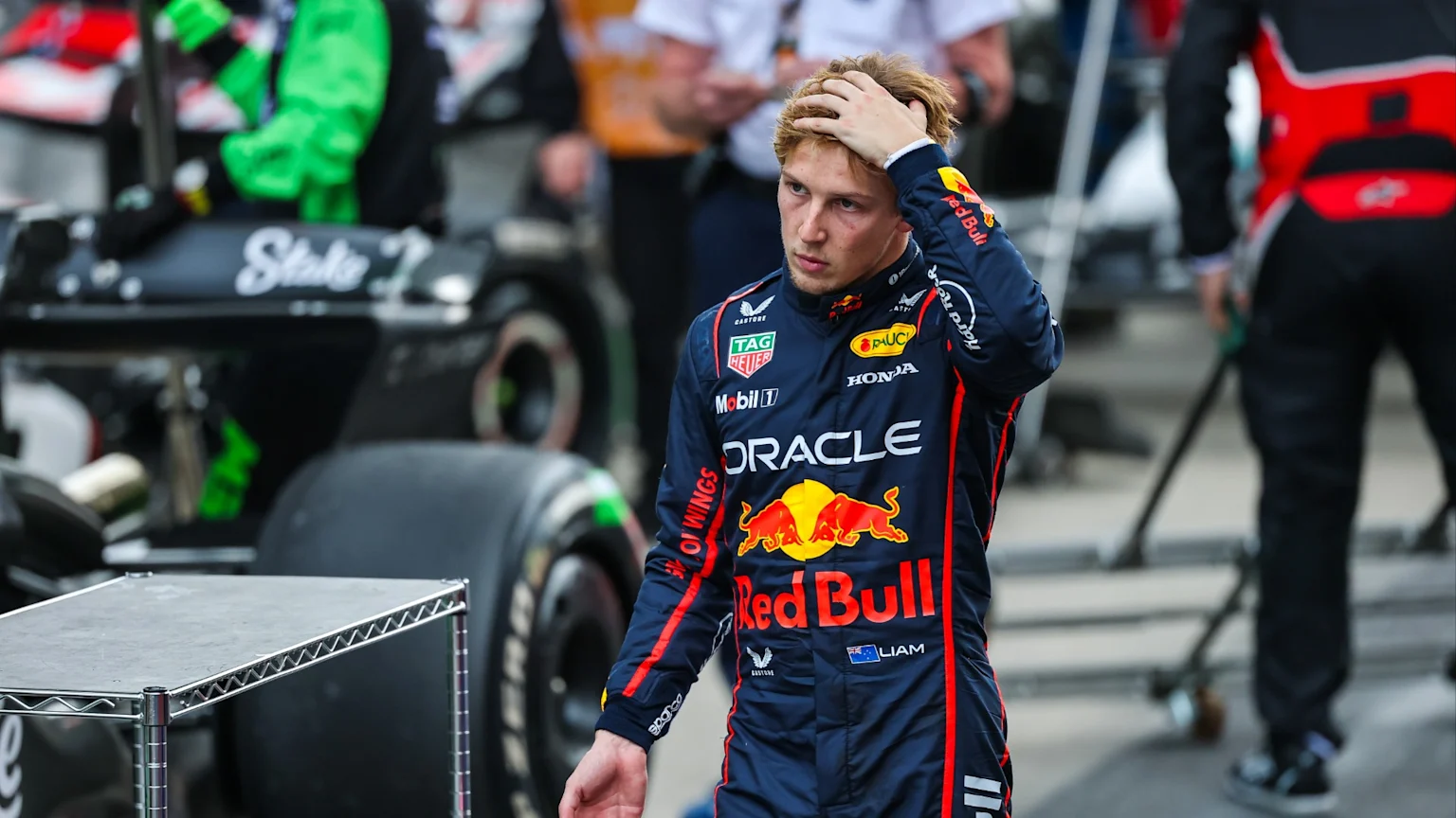 Liam Lawson, in a navy Red Bull racing suit with Red Bull and sponsor logos, stands with a hand on his head, looking serious, near a blurred black F1 car with 'Stake' text.