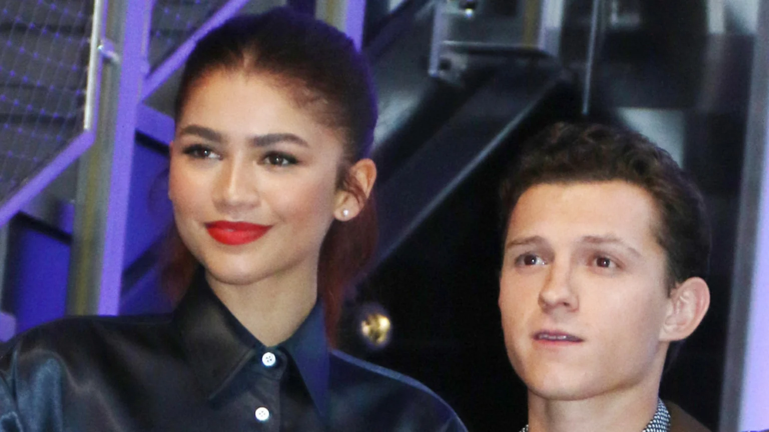 Zendaya, with dark hair and red lipstick, smiles next to Tom Holland, who looks forward, against a dark background with purple elements.