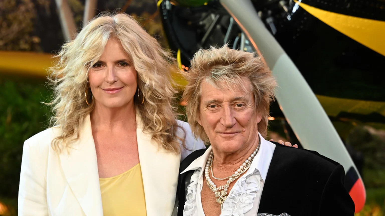Penny Lancaster, with long blonde hair and a white blazer, smiles next to Rod Stewart, who wears a ruffled shirt and necklaces, with an airplane visible behind them.