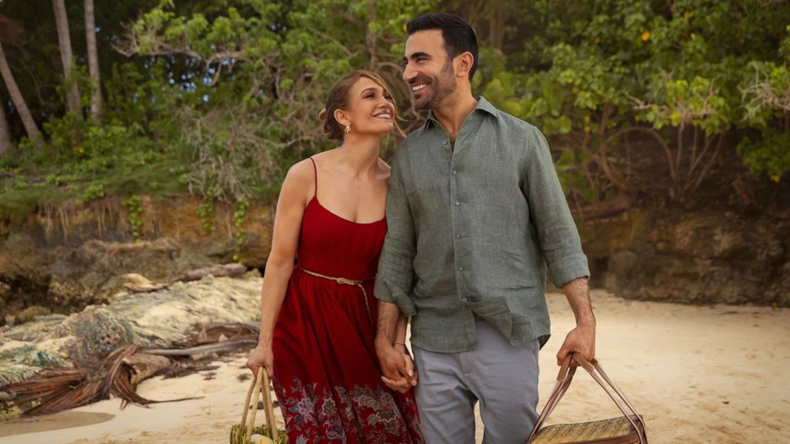 A woman in a red dress and a man in a green shirt walk hand-in-hand on a sandy beach, smiling lovingly at each other, while carrying woven bags with lush green foliage behind them.