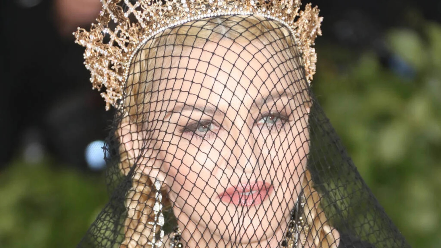 Close-up of Madonna, with blonde hair, wearing a jeweled gold crown and a black net veil over her face, revealing her eyes and lips, against a blurred dark and green background.