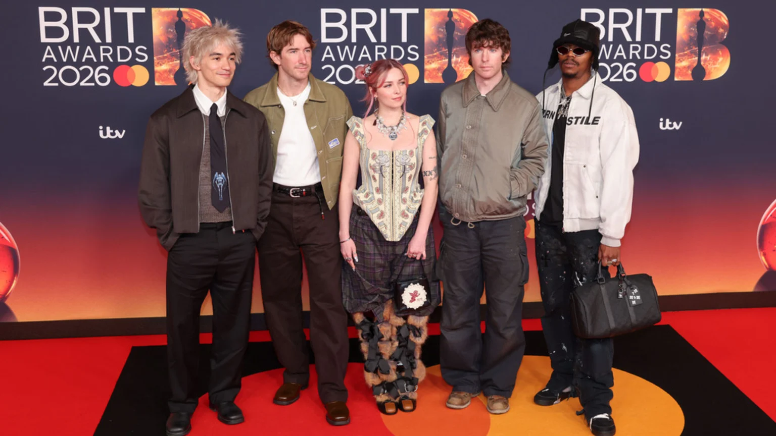 Five people, including a woman with pink hair and a man wearing a white jacket with "URN ASTILE" written on it, pose on a red carpet at the BRIT Awards 2026.