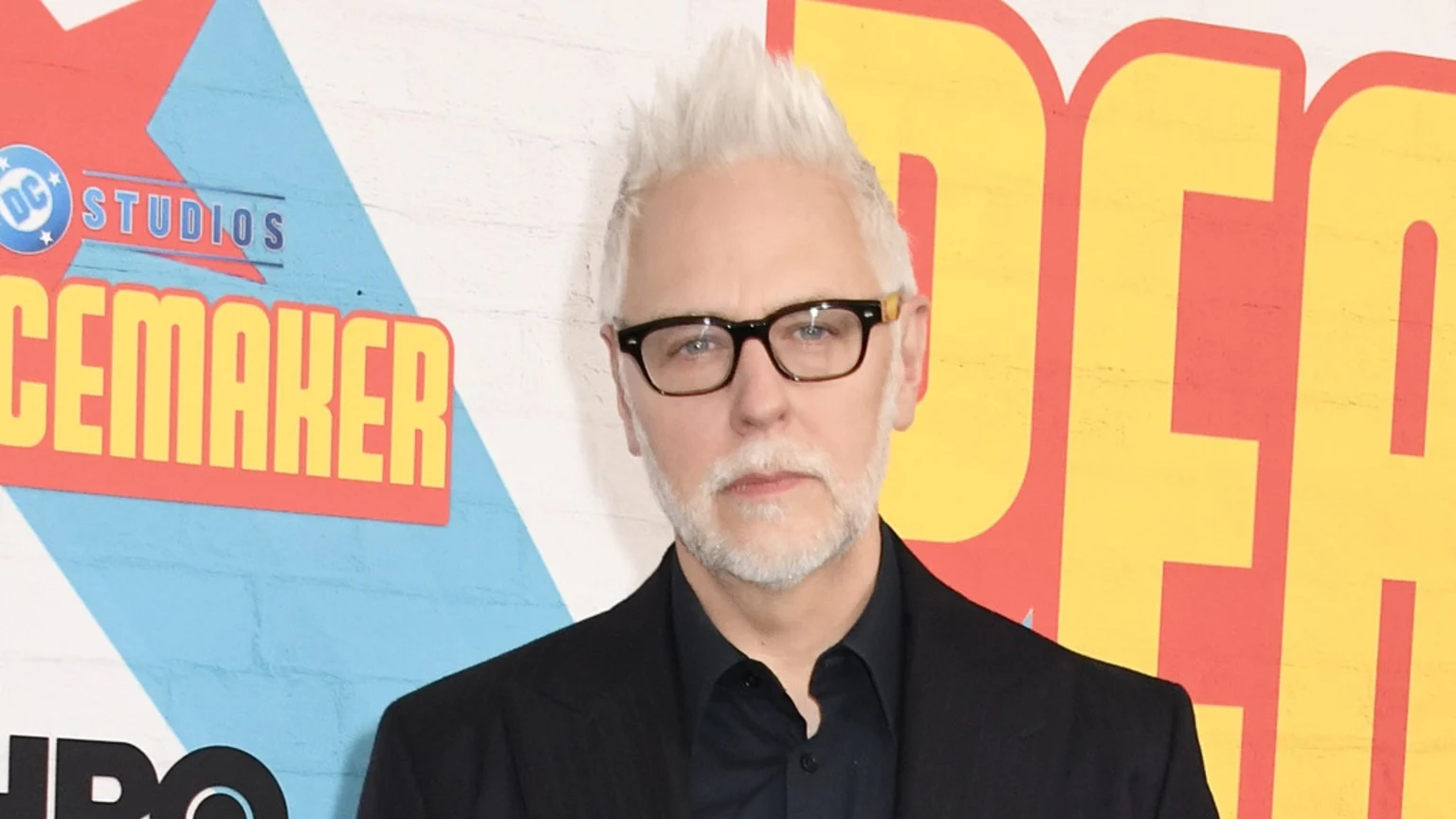 James Gunn, a man with spiky white hair, a white beard, and glasses, looks directly at the camera in front of a colorful backdrop featuring "DC Studios" and "Peacemaker" logos.