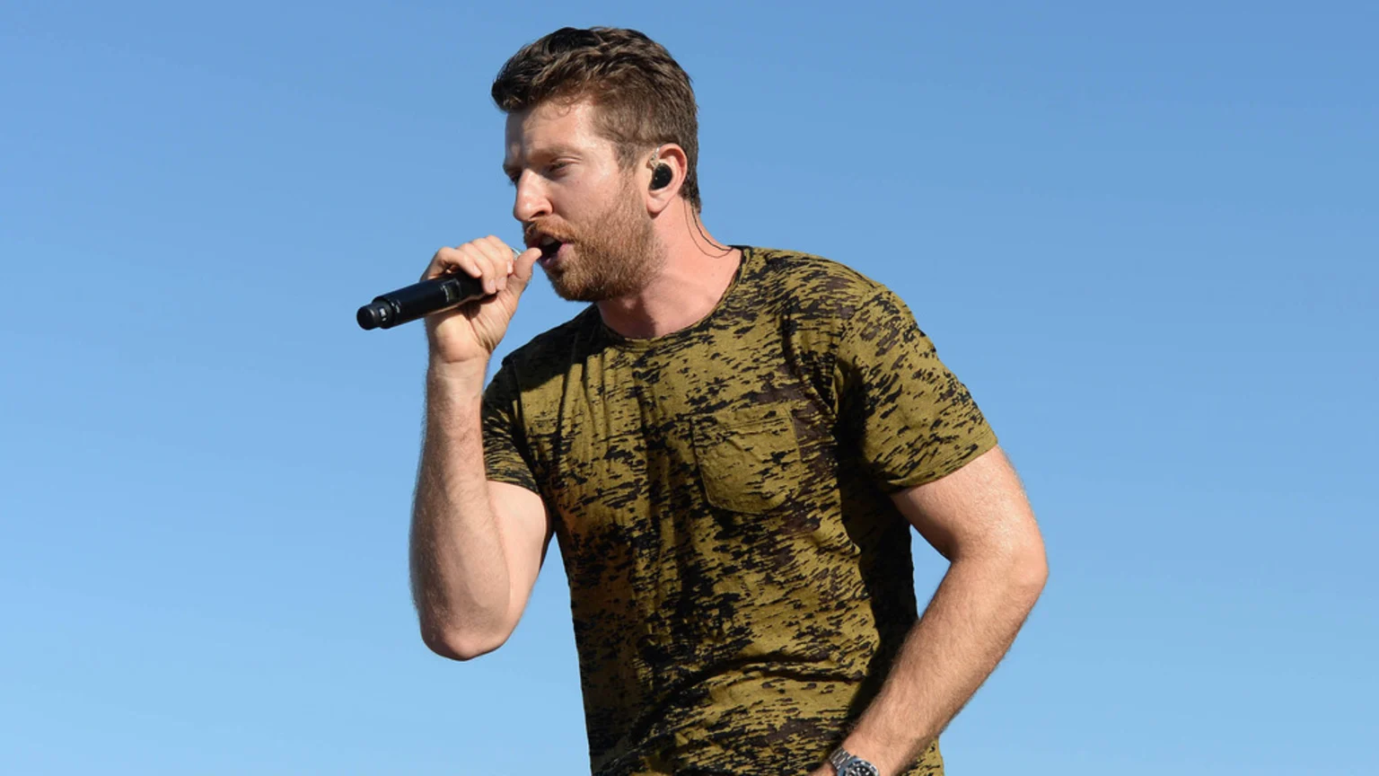 Brett Eldredge sings into a microphone outdoors against a clear blue sky, wearing a patterned olive green t-shirt and an in-ear monitor.
