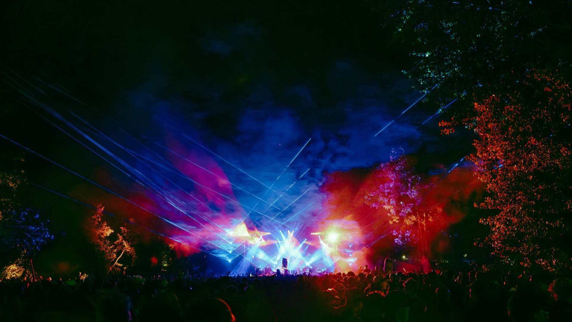 A nighttime outdoor music festival scene features a large crowd silhouetted against a brightly lit stage with vibrant blue, red, and purple lights, lasers, and smoke filling the sky and illuminating nearby trees.