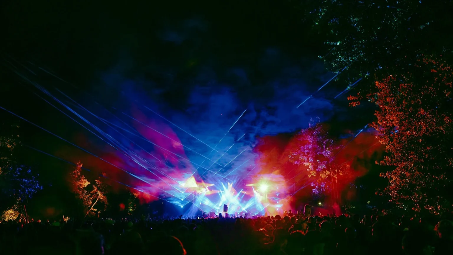 A nighttime outdoor music festival scene features a large crowd silhouetted against a brightly lit stage with vibrant blue, red, and purple lights, lasers, and smoke filling the sky and illuminating nearby trees.
