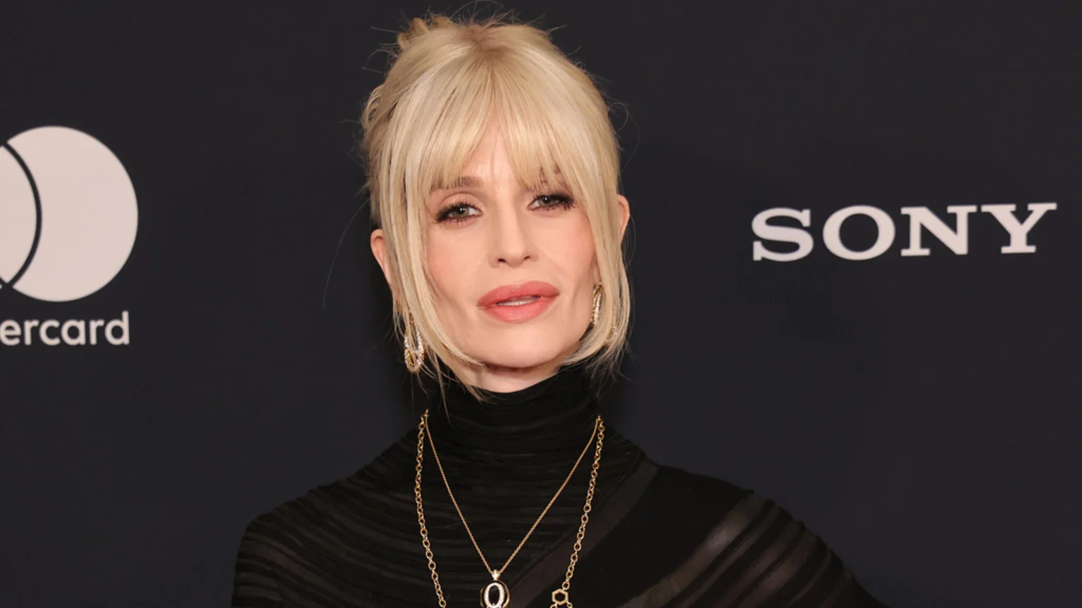 Close-up of a blonde woman with bangs, wearing a black turtleneck and gold jewelry, against a dark background with visible Mastercard and Sony logos.