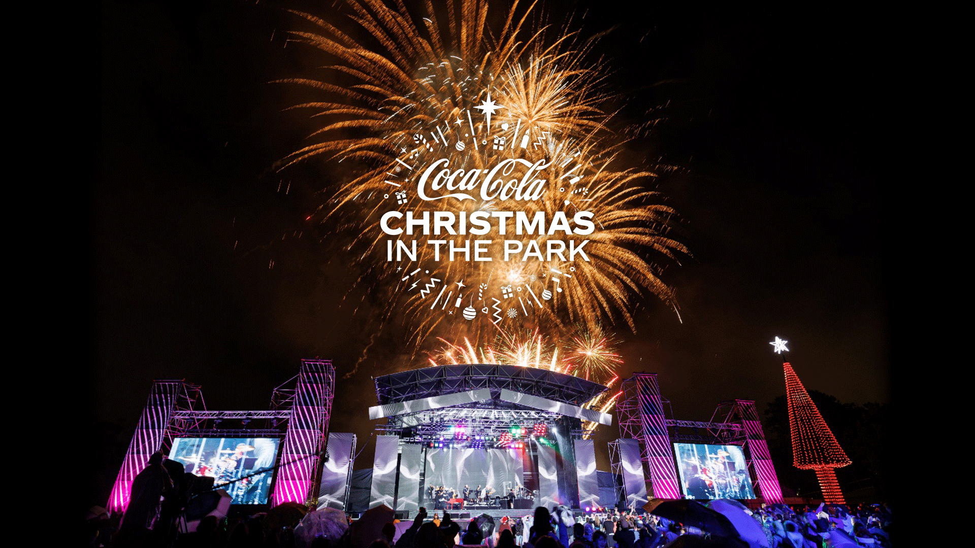 White "Coca-Cola Christmas in the Park" text, surrounded by festive icons, is set against a backdrop of golden fireworks.