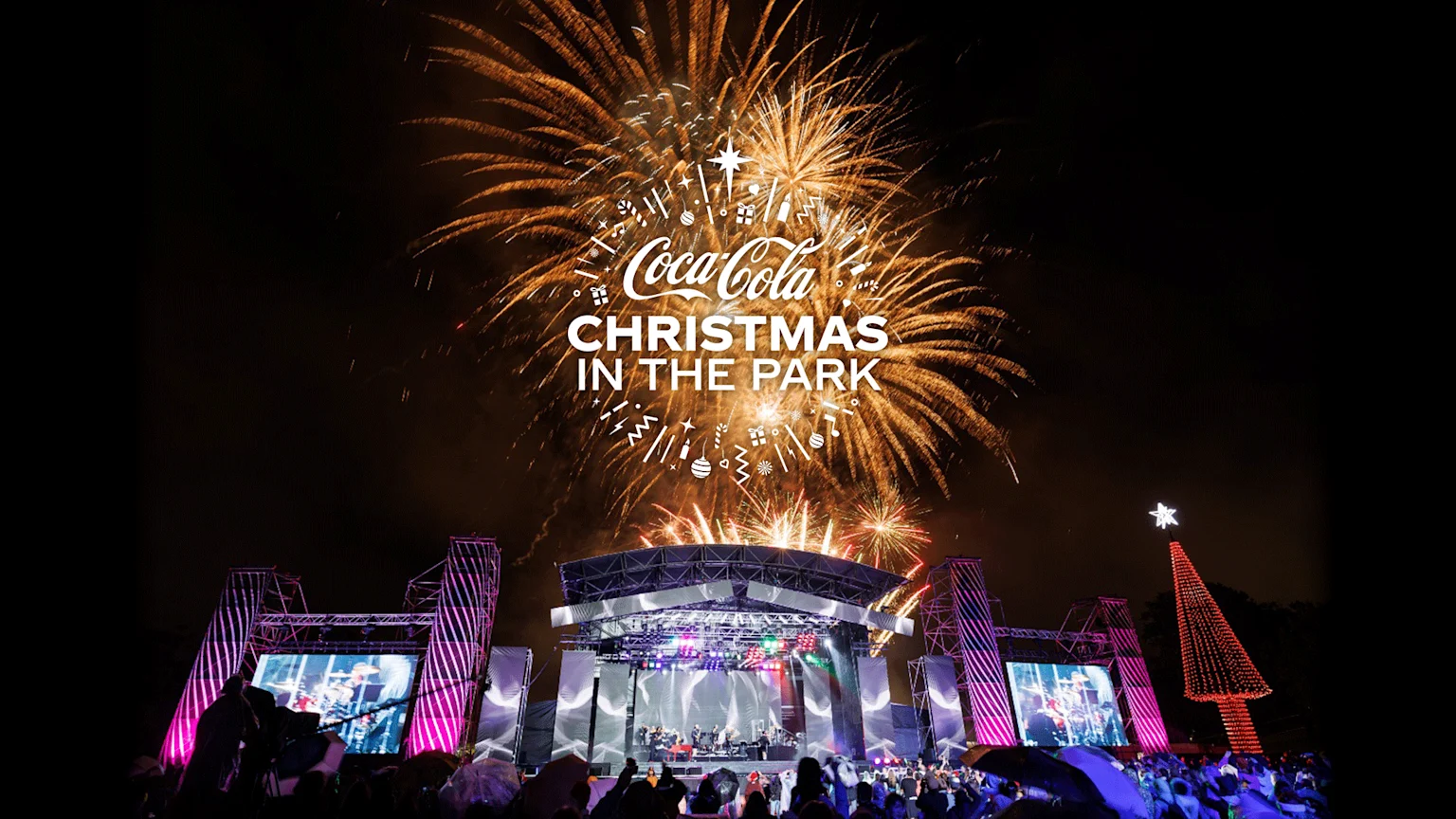White "Coca-Cola Christmas in the Park" text, surrounded by festive icons, is set against a backdrop of golden fireworks.