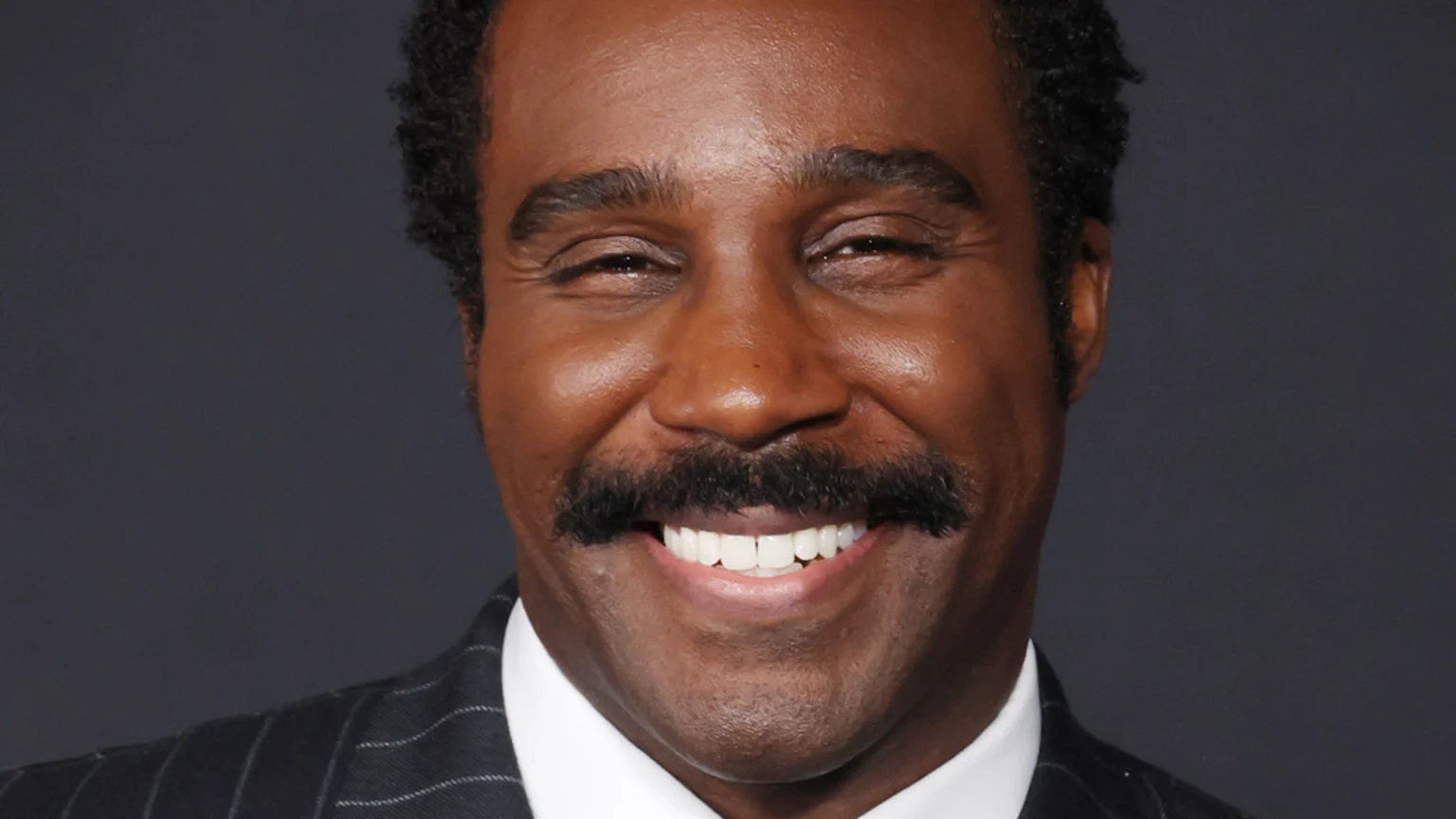 A close-up of a smiling Black man with a mustache and short curly hair, wearing a dark pinstripe suit and white shirt, against a dark background.