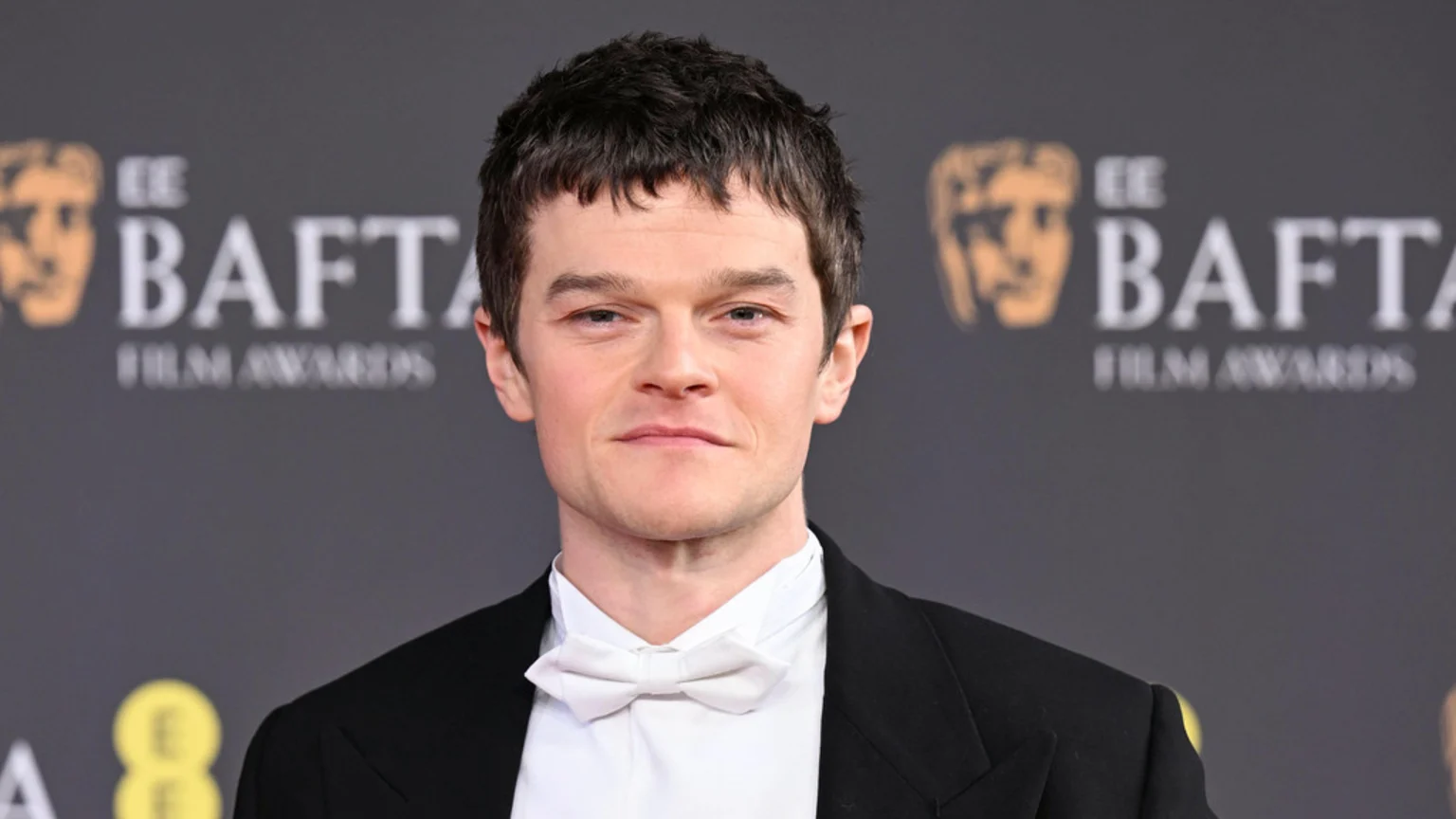Robert Aramayo in a black suit, white shirt, and white bow tie, looking directly at the camera with a subtle smile against a blurred EE BAFTA Film Awards background.