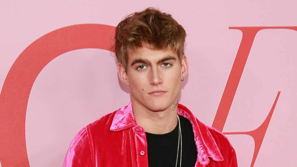 A man with light brown wavy hair, a mole below his left eye, and a neck tattoo wears a shiny hot pink jacket and a silver necklace, looking at the camera against a pink background with large red abstract shapes.