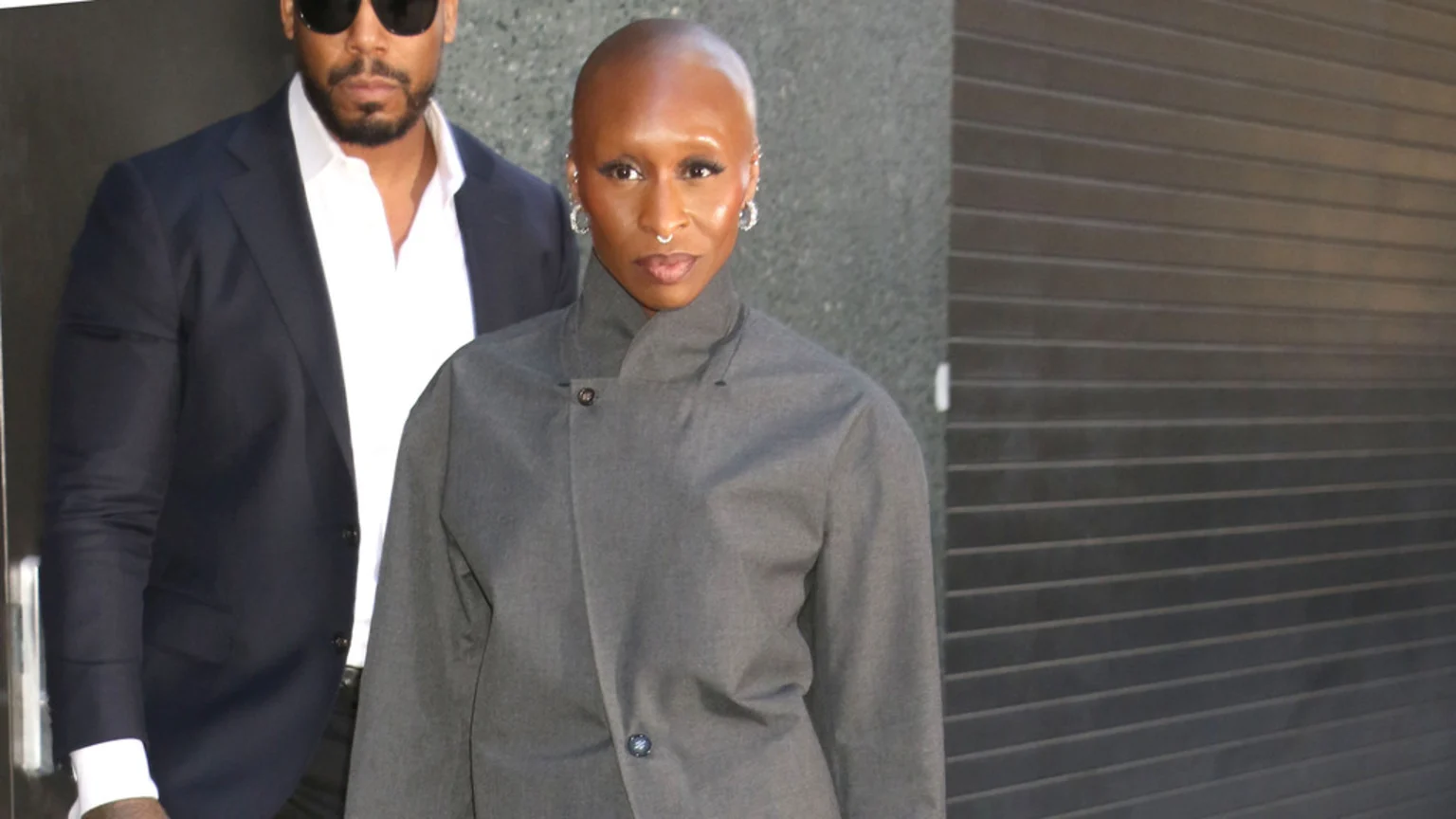 Cynthia Erivo, a dark-skinned person with a shaved head, wearing a grey jacket, nose ring, and earrings, stands in front of a dark, striped background, with a man in sunglasses and a dark suit partially visible behind her.