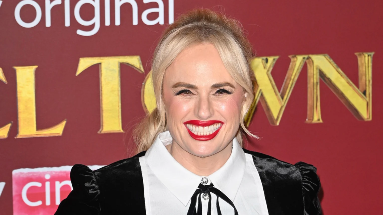 Rebel Wilson, a blonde woman, smiles broadly with bright red lipstick, wearing a black velvet dress with a white collar and black tie detail, against a red background with gold lettering.