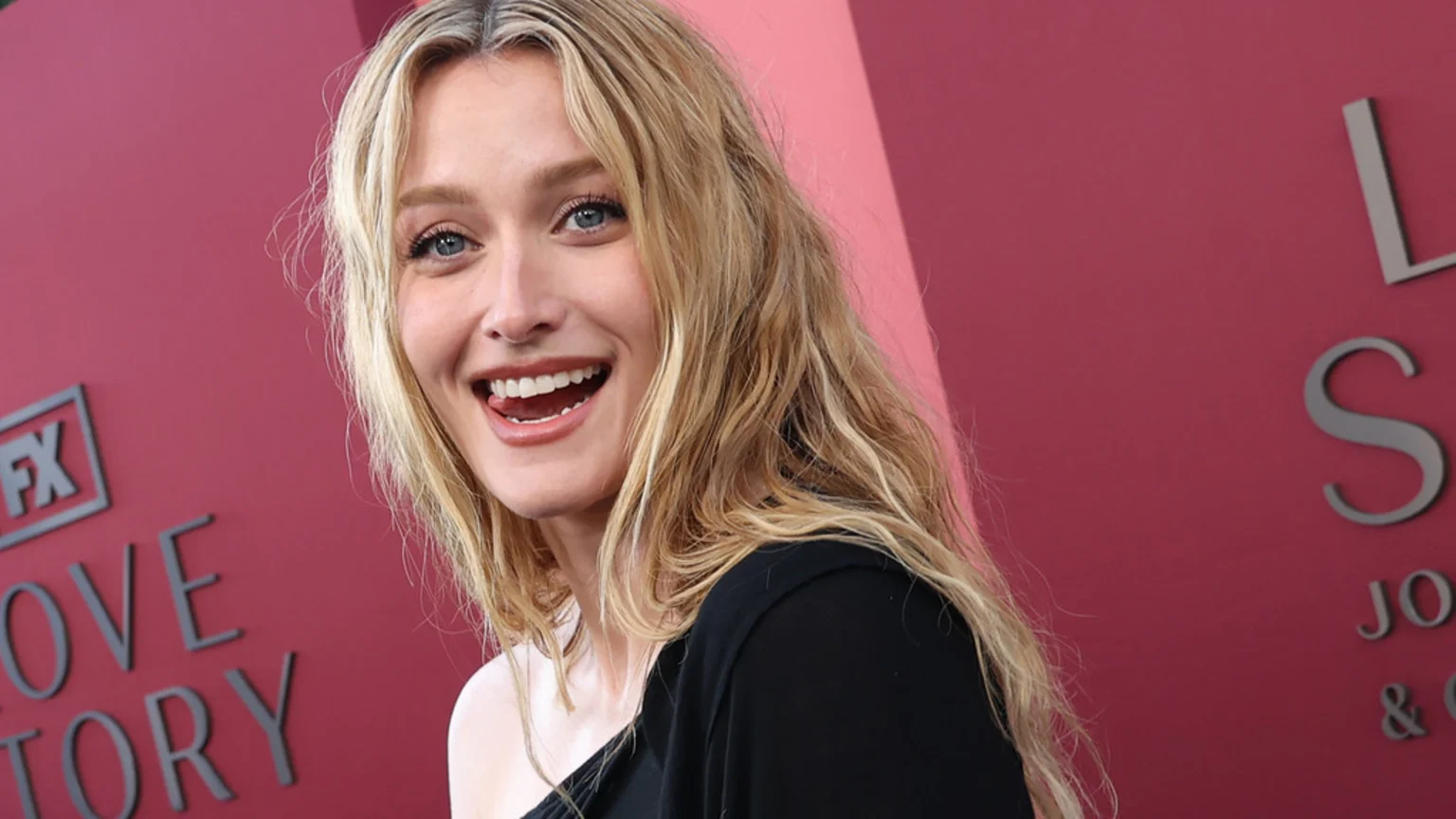A blonde woman with blue eyes, wearing a black top, smiles widely with her tongue slightly out, posing against a burgundy background with grey text.