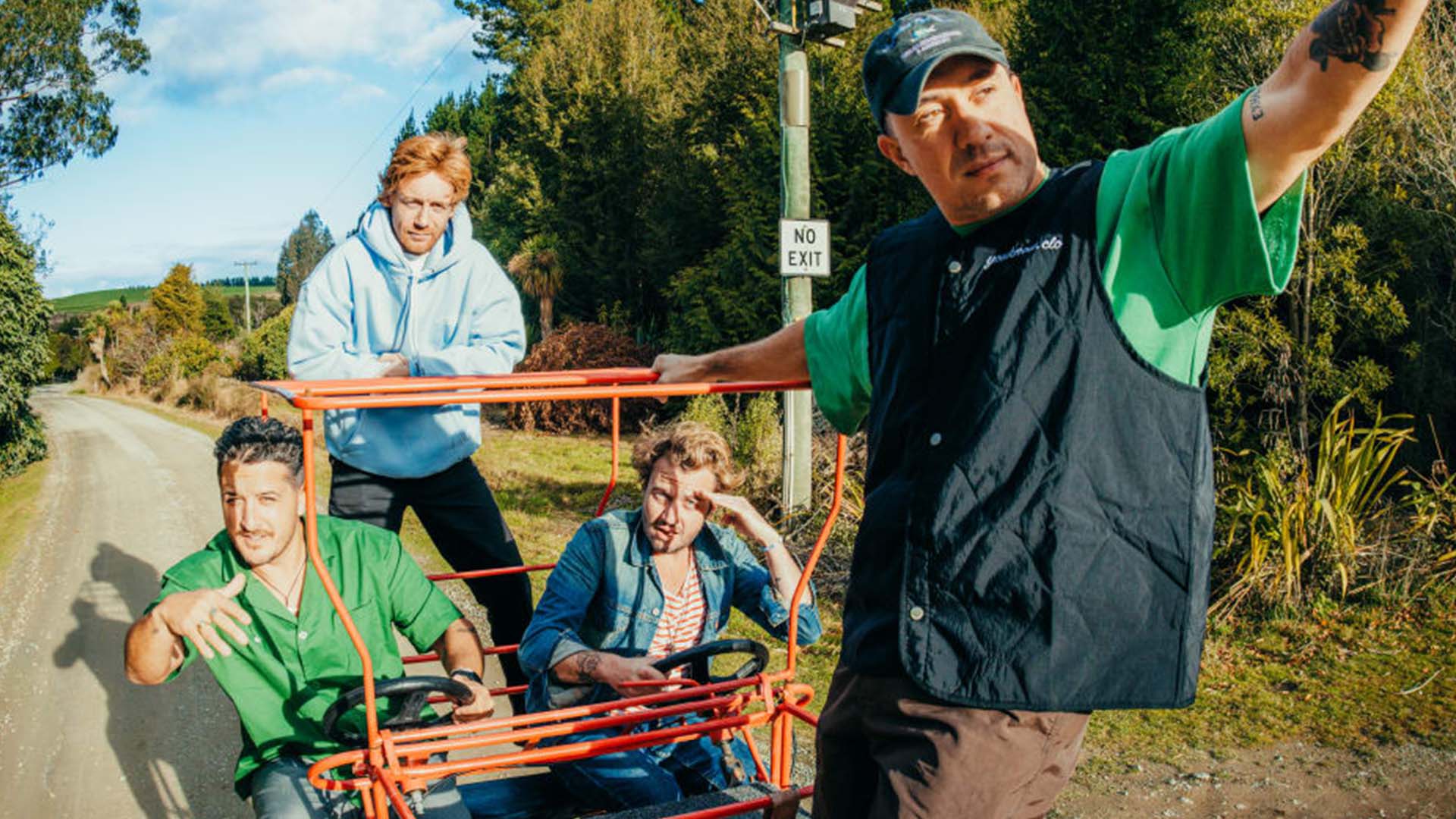A group of four men pose with a red multi-person pedal vehicle on a rural dirt road, surrounded by trees and a "NO EXIT" sign.