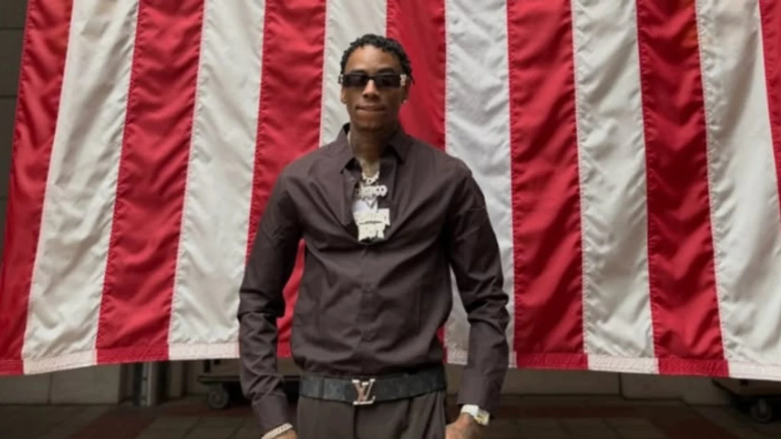 A man wearing sunglasses, a dark brown collared shirt, and a large chain stands centered in front of a red and white striped flag.