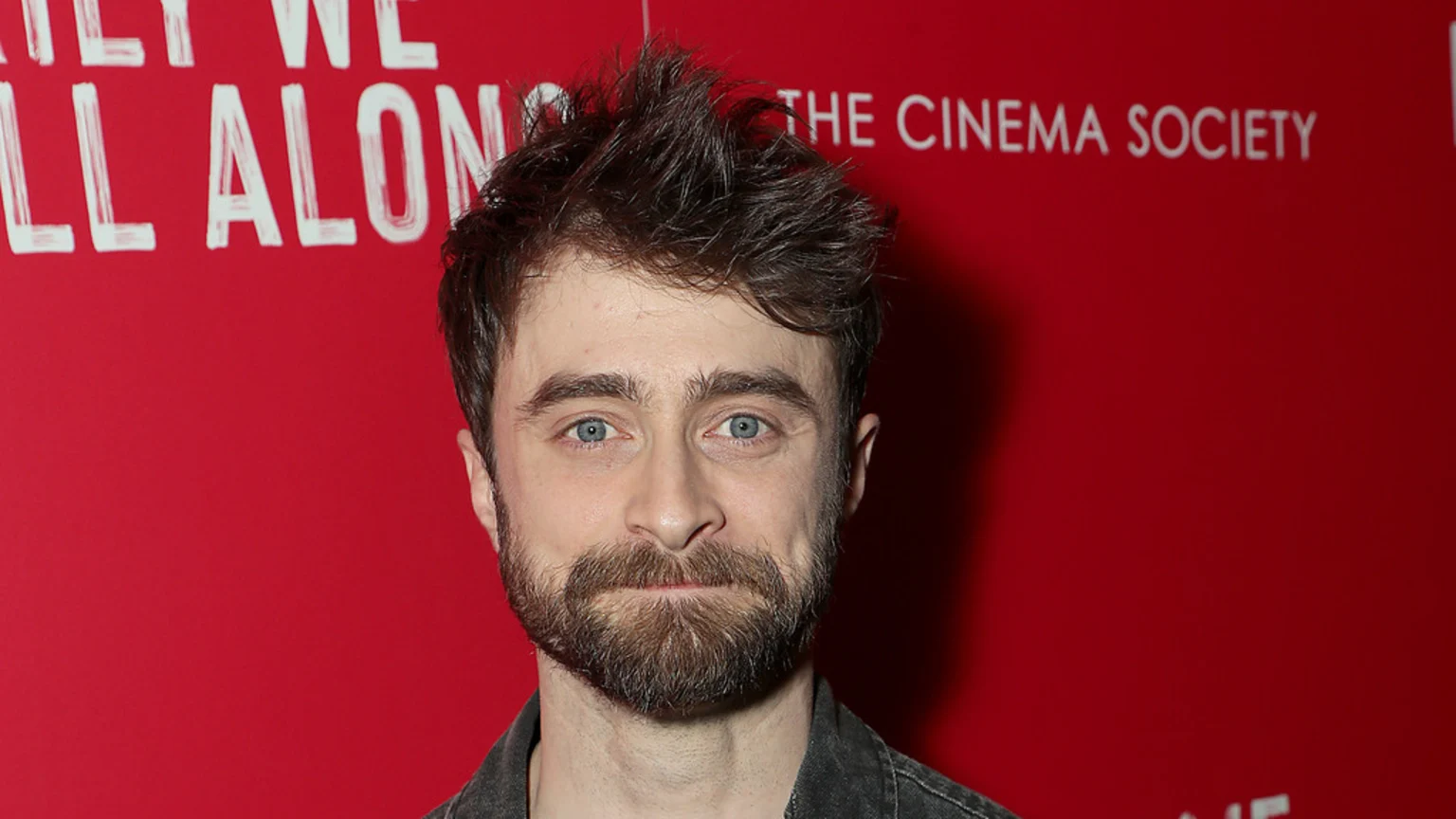 Daniel Radcliffe, with blue eyes, a beard, and messy brown hair, looks directly at the camera against a red background with partially visible white text.