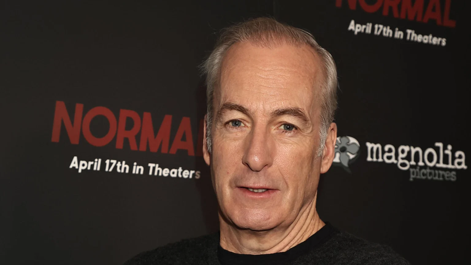 A man with grey hair and blue eyes looks directly at the camera, standing in front of a dark backdrop with the red word "NORMAL," "April 17th in Theaters," and a "magnolia pictures" logo.