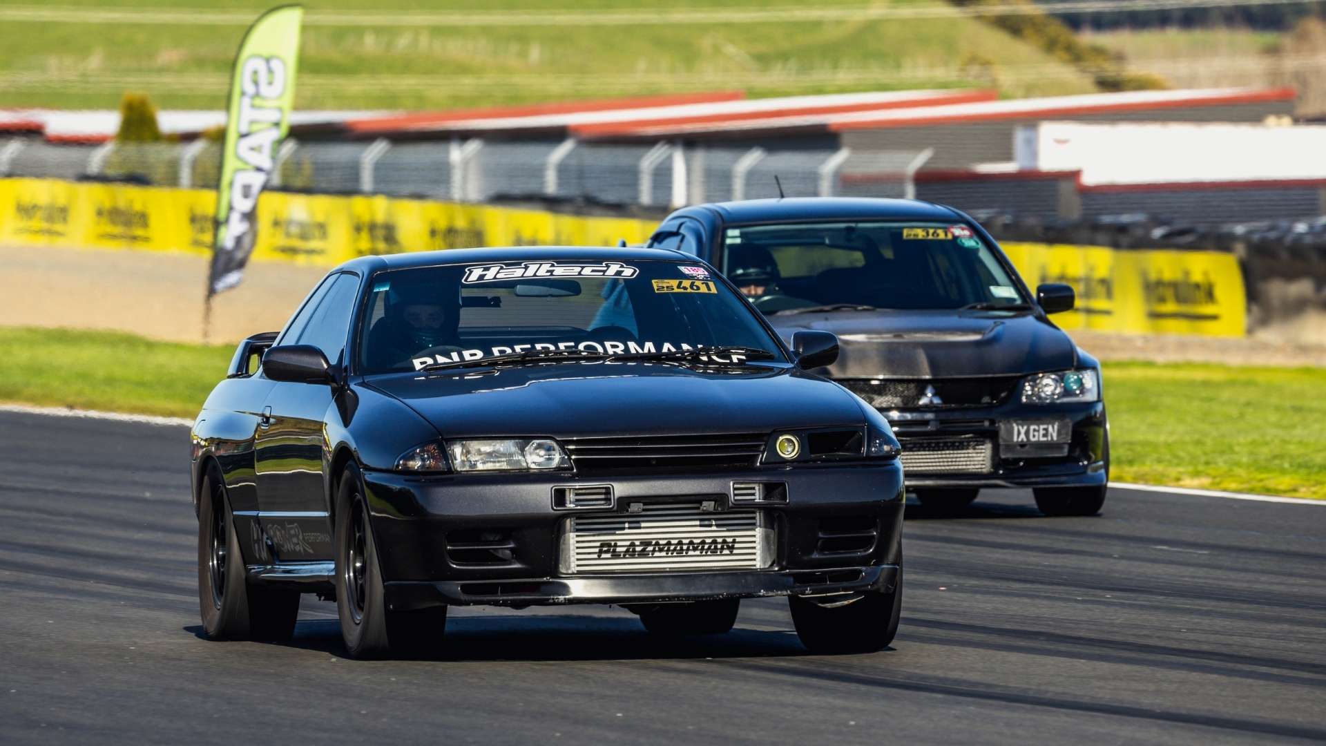 A black Nissan Skyline R32 race car with "Haltech" on its windshield and "PLAZMAMAN" on its intercooler, followed by a dark Mitsubishi Lancer Evolution, on a race track.
