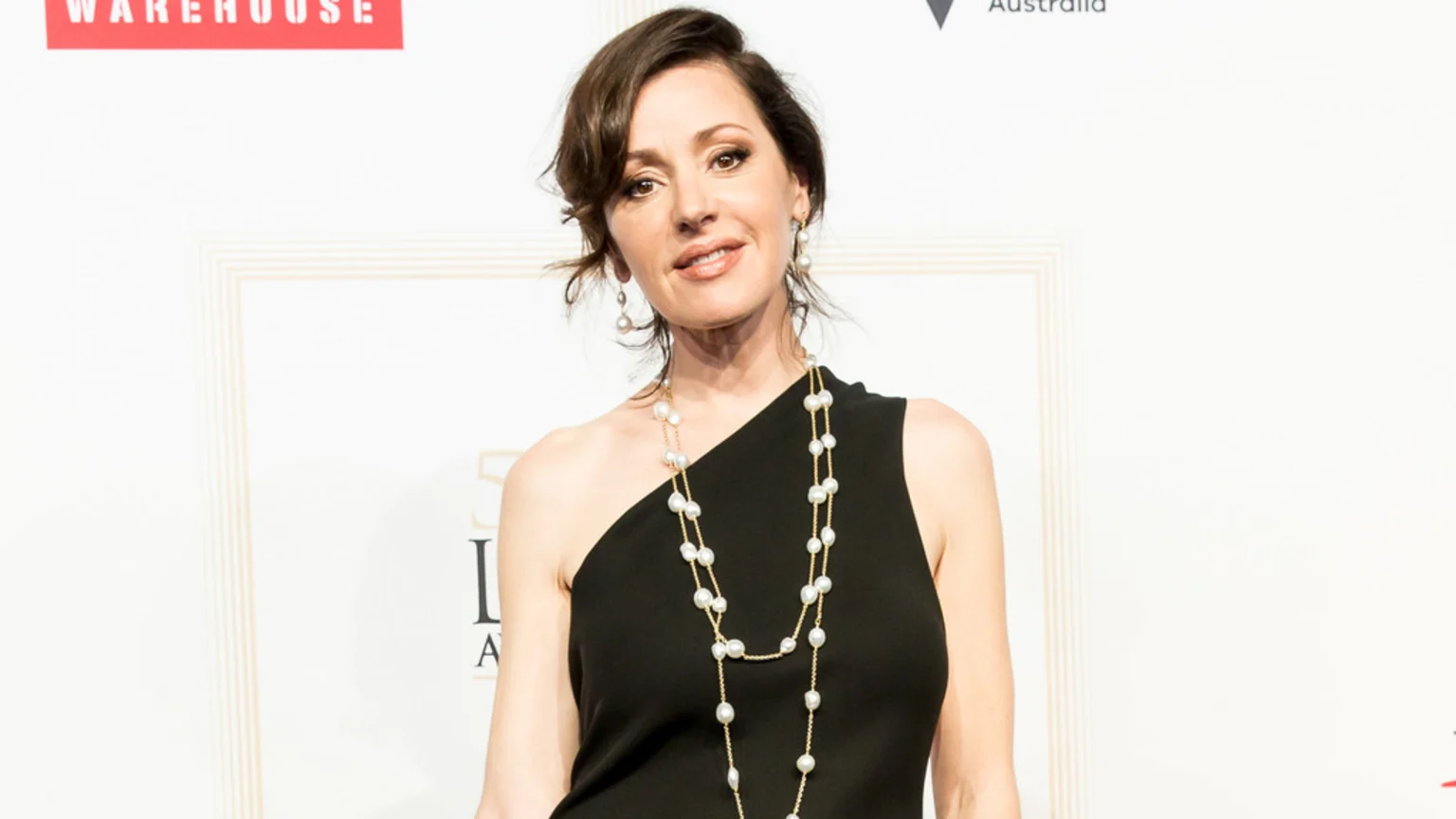 Tina Arena smiles and looks at the camera, wearing a black one-shoulder top, a long pearl necklace, and pearl earrings against a light background.