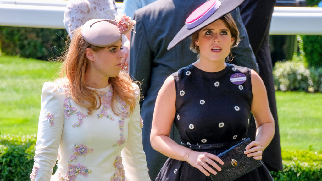 Princesses Beatrice and Eugenie walk side-by-side outdoors. Beatrice wears a cream dress with colorful floral embellishments and a fascinator, while Eugenie wears a black dress with white floral patterns, a wide-brimmed hat, and holds a clutch.