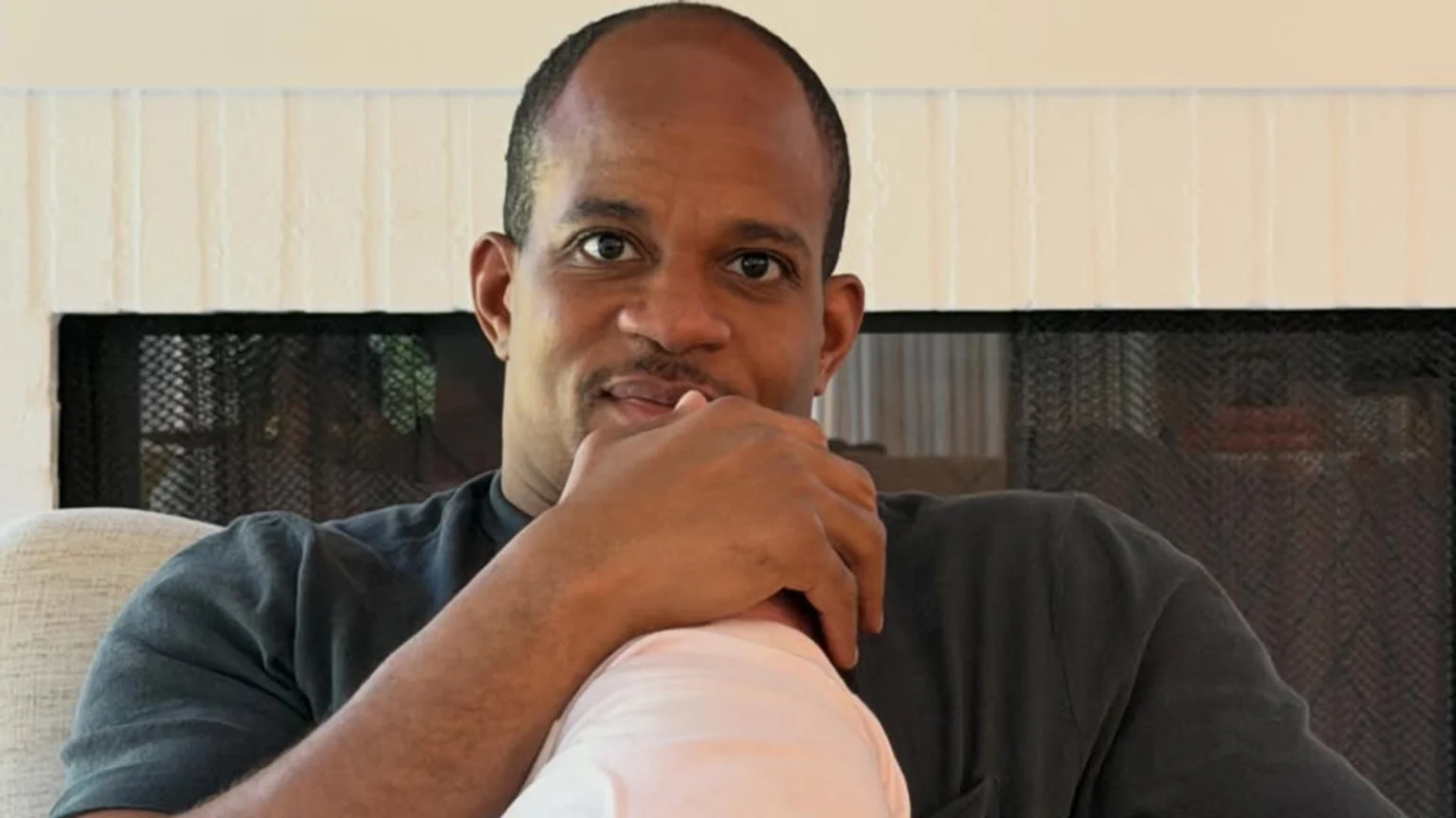 A dark-skinned man with short hair and a mustache looks at the camera, holding his hand to his chin while cradling a baby wearing a light-colored outfit.