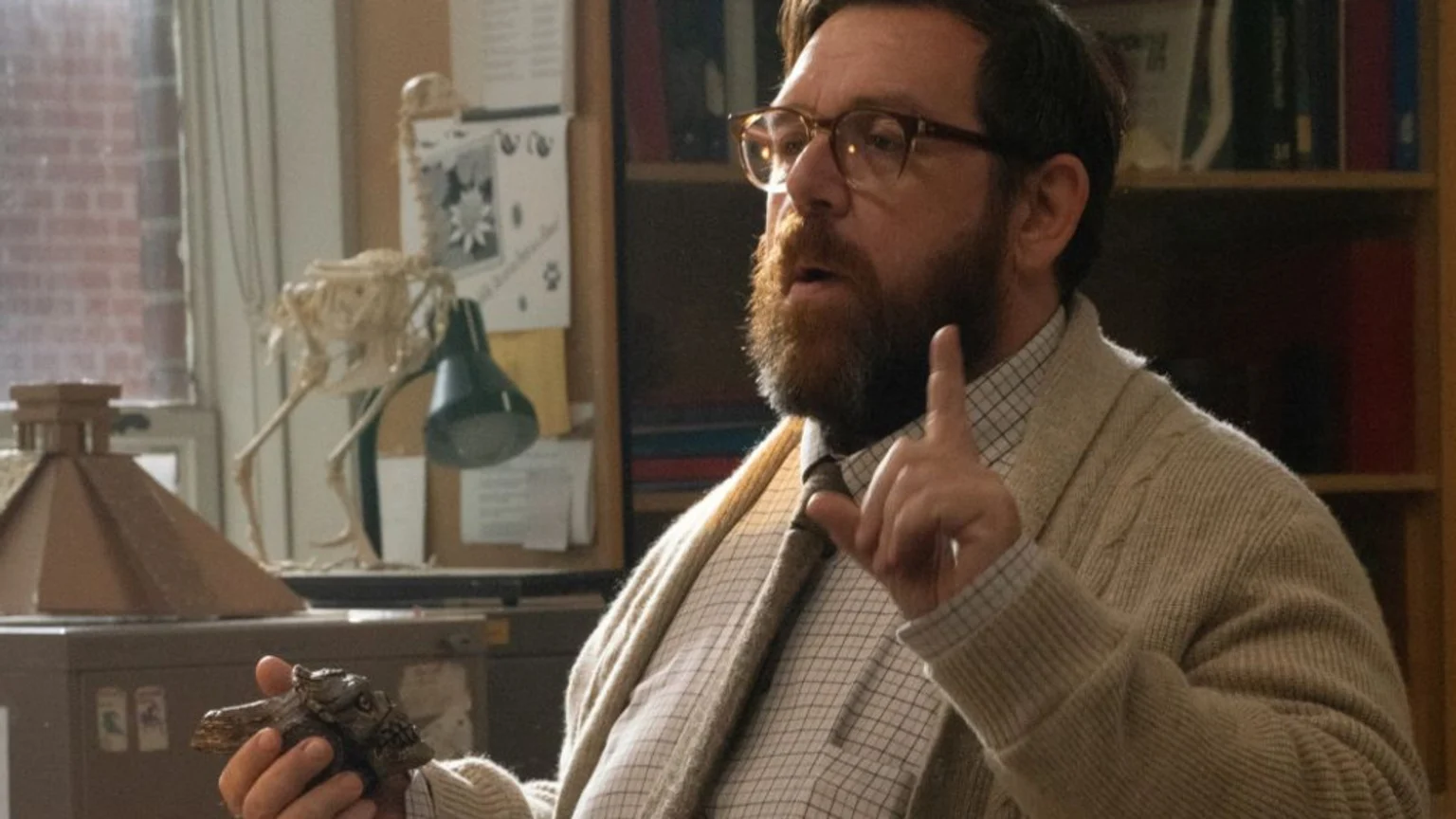 Close-up of a bearded man wearing glasses, a patterned shirt, tie, and cable-knit cardigan, speaking and gesturing with his right index finger raised. He holds a small, dark, textured object in his left hand. Background includes a bird skeleton scientific display, desk lamp, bookshelf with books and posters, a window showing a red brick building, and other office items.