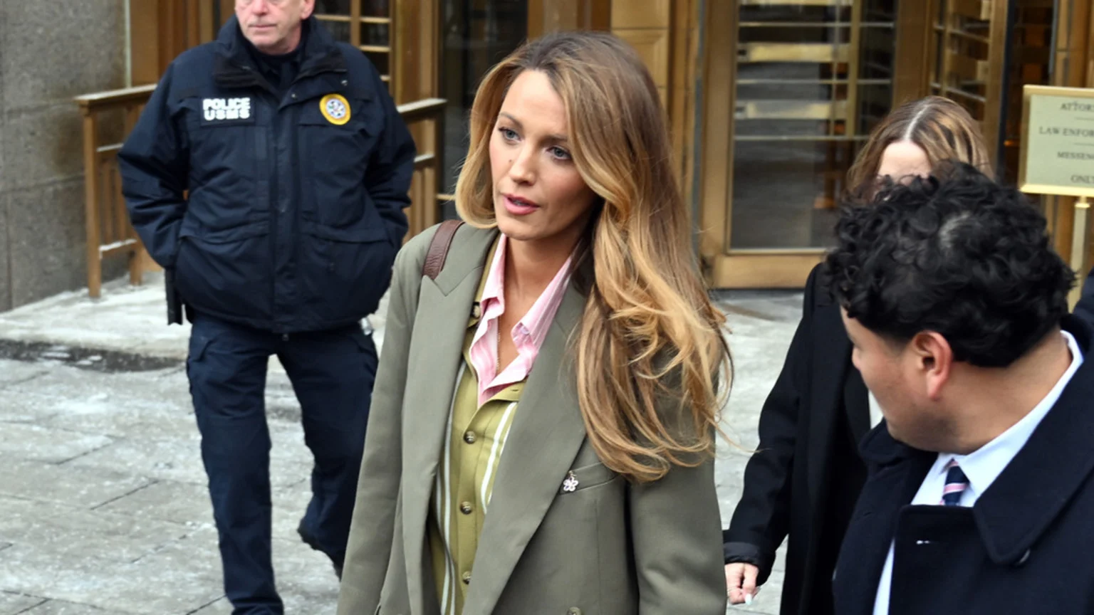 Blake Lively, with long blonde hair, wears a green blazer and striped shirt, walking past a police officer in a dark uniform and other individuals outdoors.