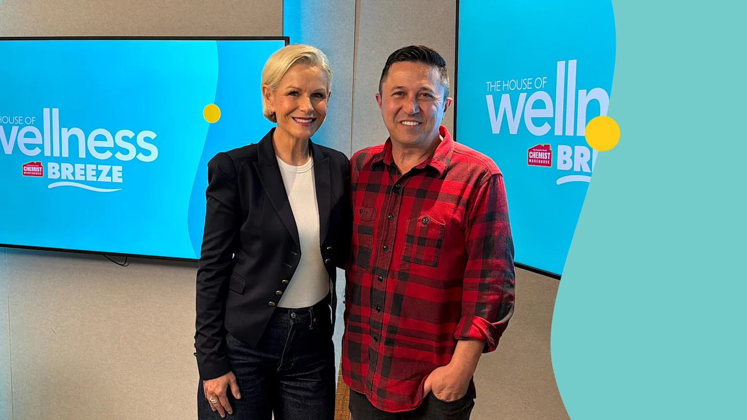 Lorraine Downes, with short blonde hair and a dark blazer, smiles next to a man in a red plaid shirt, with "The House of Wellness" screens behind them.