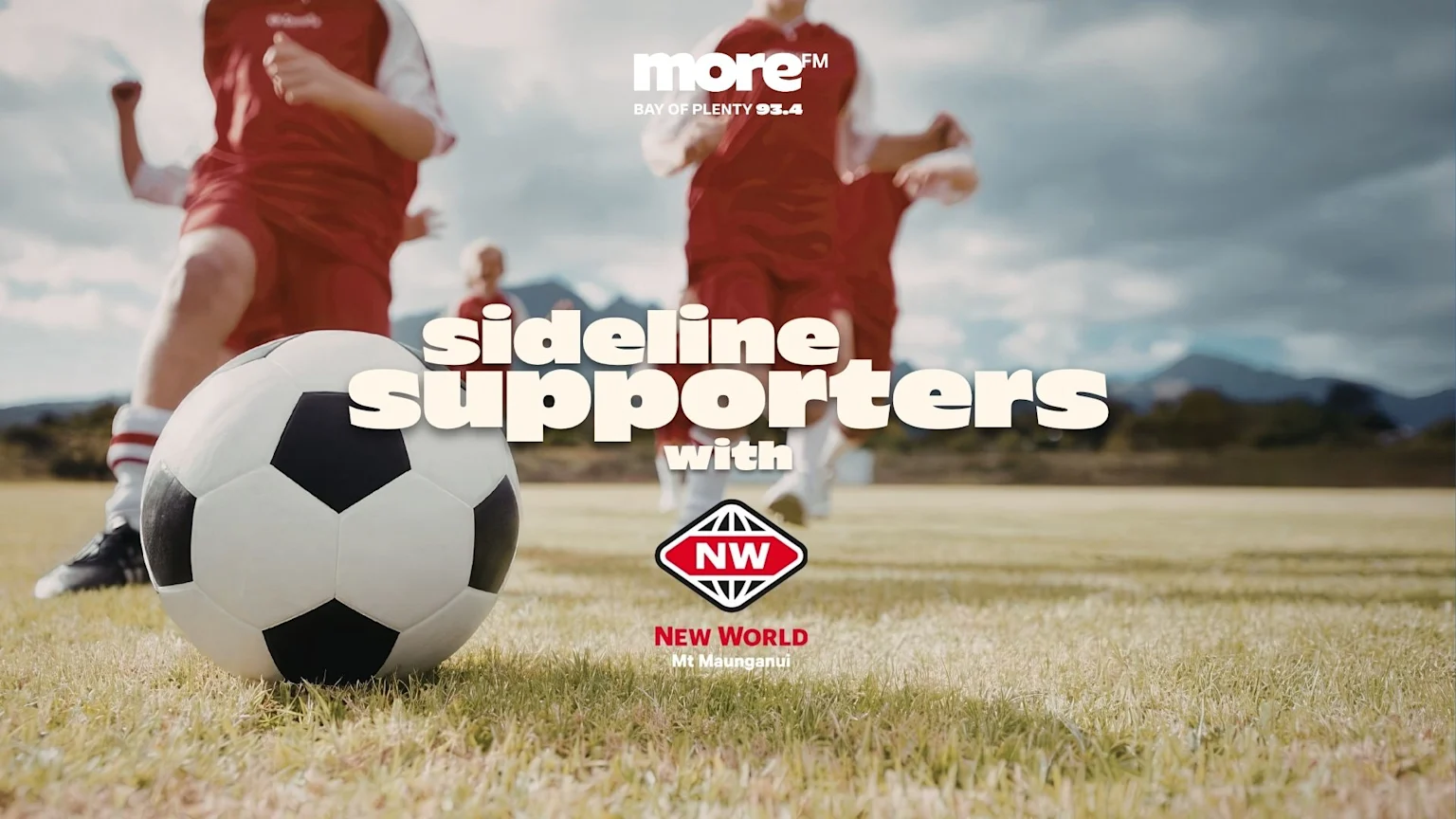 A soccer ball on a grassy field with blurred figures of players in red jerseys and mountains, featuring text "sideline supporters with", "more FM", and "NEW WORLD Mt Maunganui" logos.
