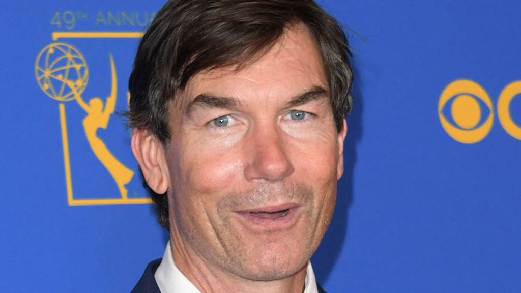 Close-up of a smiling man with dark hair, blue eyes, and light stubble, wearing a white shirt, against a blue background with partial Emmy and CBS logos.